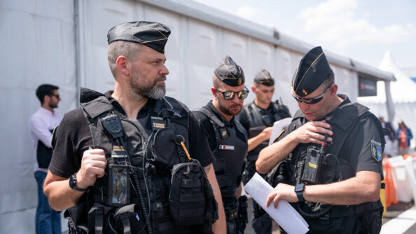 Trois gendarmes mobiles: celui de gauche de profil, celui du milieu de face, celui de droit de face en train de parler dans la radio accrochée à sa chasuble, il tient une feuille blanche pliée dans la main.