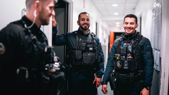 Trois gendarmes souriant en tenue d'intervention, en intérieur dans un couloir éclairé.