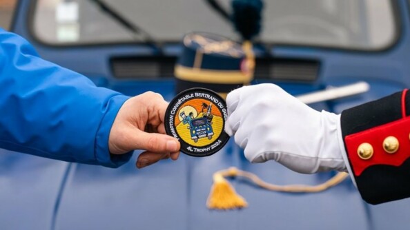 Devant le capot bleu marine d'une Renaud 4L, deux mains tendues de part et d'autre du capot, tiennent chacune le bord d'un &eacute;cusson comportant la mention "promotion Conn&eacute;table Bertrand du Guesclin - 4L Trophy 2024". A gauche de l'&eacute;cusson, l'avant-bras est v&ecirc;tu d'un v&ecirc;tement bleu moyen, tandis qu'&agrave; droite, il s'agit de la manche d'un uniforme bleu marine et rouge avec des boutons dor&eacute;s. La main qui tient l'&eacute;cusson rev&ecirc;t un gant blanc.