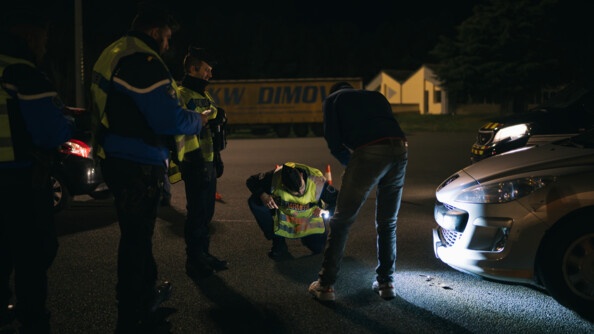 Trois gendarmes faisant un contrôle routier, dont un accroupi. Il regarde le dessous d'une voiture avec une lampe torche. Le conducteur de la voiture est à côté d'eux, de dos.
