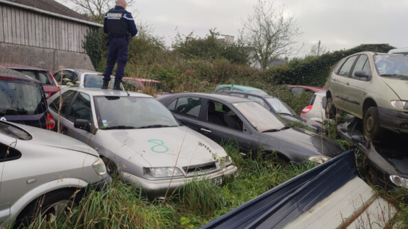 Un gendarme de dos, debout sur le toit d'une épave de voiture, au milieu d'une casse automobile dans un champ.