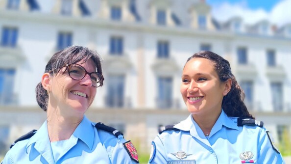 Deux femmes gendarmes en uniforme, se tiennent côte à côte, souriantes, devant un bâtiment ancien de couleur clair, qui apparait flouté en arrière plan