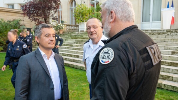 Gérald Darmanin se tient debout, dans les jardins de l'hôtel de Beauvau, devant de larges escaliers, où il discute avec un homme en uniforme noir, sur lequel est inscrite la mention RAID. A leurs côtes se trouve le directeur général de la gendarmerie nationale.