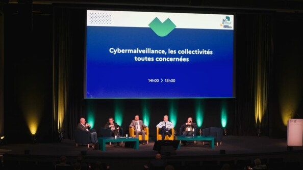 Dans un amphithéâtre plongé dans l'obscurité, excepté des lumières vertes et jaunes au fond venant du sol, une table ronde. Sur l'estrade noire, de part et d'autres, deux tables vertes. Conq hommes assis avec des micros, celui de gauche s'exprime. Deuxième en partant de la droite, le général Boget, commandant le ComCyberGend. En arrière-plan, écrit en blanc sur fond bleu roi, la thématique de la table ronde: "Cybermalveillance, les collectivités toutes concernées"