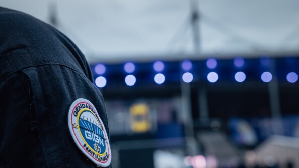 Sur l'épaule d'un gendarme en uniforme, apparait un écusson sur lequel est inscrit : Gendarmerie nationale - GIGN. On aperçoit en arrière plan les lumières floutées du Stade de France