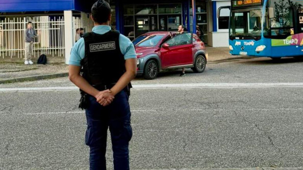 Un gendarme de dos sur la route , devant un lycée Jean Monnet, au pied duquel se trouve une voiture rouge où monte un homme et derrière elle un bus bleu