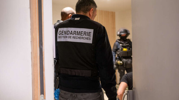 Un homme de dos, dans l'encadrement d'une porte. Il est vêtu d'un jean anthracite, d'une veste noire et d'une chasuble noire portant la mention "GENDARMERIE SECTION DE RECHERCHES" écrite en noir dans un rectangle blanc au milieu du dos. Au second plan, à gauche, on aperçoit la tête d'un homme de profil, et en bas, à droite, le bras et l'épaule d'un homme vu de dos, en T-shirt noir, que l'on imagine assis et menotté. En arrière-plan, à droite, au fond de la pièce, un gendarme mobile en tenue d'intervention, portant protections individuelles et casque.