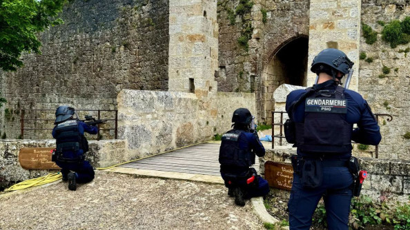 Trois gendarmes équipés en casque et gilet lourd devant un pont levis, positionnés debout ou un genoux en terre
