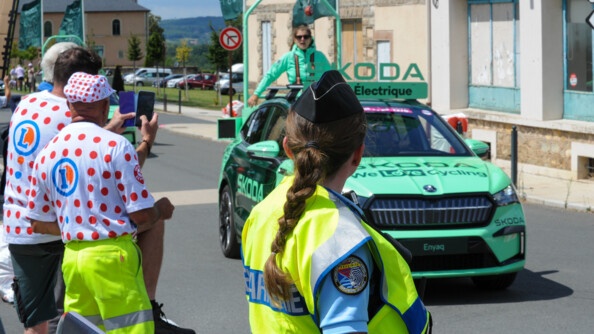 Le long de la route traversée par un véhicule de la caravane Skoda, des badauds et une gendarme de dos avec chasuble jaune