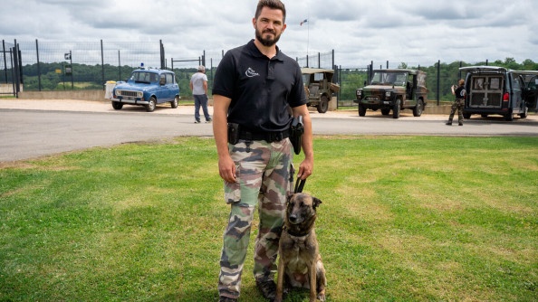 Photo d'un militaire en treillis kaki, avec un polo noir, tenant en laisse en berger belge malinois assis à ses côtés