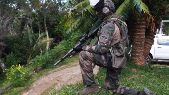 Un militaire en tenue de camouflage est accroupi, genou posé à terre, dans un paysage de jungle. Il est casque et masqué, et tient dans ses mains son fusil
