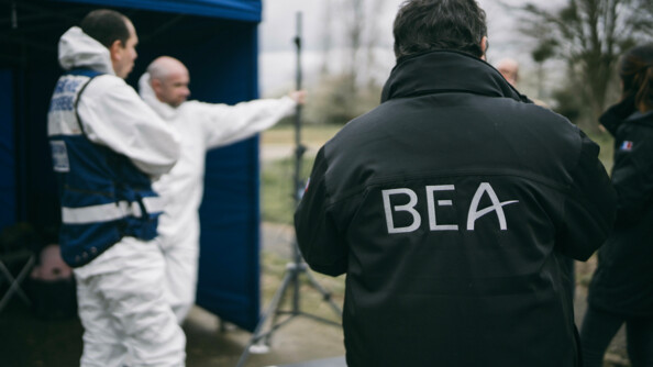 De dos, un membre du BEA, au loin floutté, deux membres des TICS dans leurs combinaisons d'intervention.