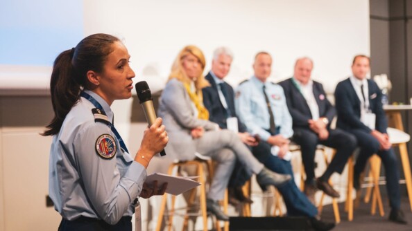 A gauche de l'image la porte-aparole de la gendarmerie nationale, le colonel Marie-Laure Pezant, de profil et tenant un micro dans lequel elle parle. Sur la partie droite, flou et en arrière-plan, une femme et cinq hommes assis sur des tabourets hauts