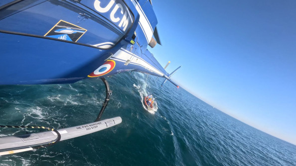 Au premier plan, la queue et le patin d'un h&eacute;licopt&egrave;re bleu de la Gendarmerie nationale en vol au-dessus de la mer, sous un grand ciel bleu sans nuage. Au-dessous de l'appareil, une vedette progresse dans la m&ecirc;me direction.