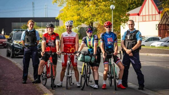 Une jeune cycliste, dans la rue, au centre de la photo, pose debout les deux mains posées sur le guidon, entourée sur sa droite par trois personnes (deux hommes cyclistes et une femme gendarme), et par deux hommes sur sa gauche (un homme cycliste et un gendarme). Derrière eux, se trouve une voiture de gendarmerie.