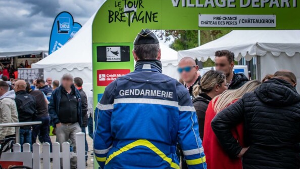 Gendarme vu de dos, au milieu du public, face au kakémono du village de départ.