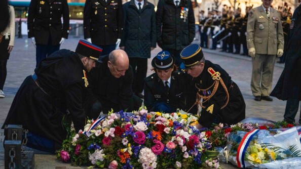 Au premier plan, un homme v&ecirc;tu de noir et trois gendarmes en grande tenue sont agenouill&eacute;s en train de d&eacute;poser une immense gerbe de fleurs sous l'Arc de Triomphe. Au second plan, cinq personnes, quatre autorit&eacute;s militaires et une civile, au garde-&agrave;-vous !