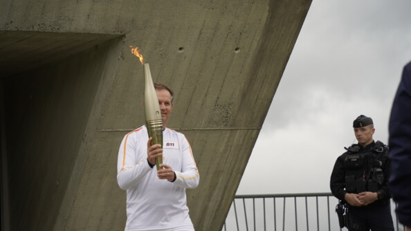 Un homme vêtu de blanc porte la flamme olympique en sortant d'un batiment de béton. A droite, un gendarme mobile