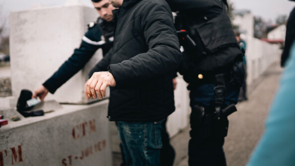 Un gendarme, entouré d'un autre gendarme, réalise une fouille sur un homme vêtu d'un jean et d'un blouson noir, se tenant debout, de biais par rapport à l'objectif.La scène se déroule sur un trottoir, par temps gris.