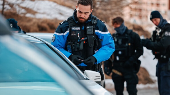 Un gendarme effectue un contrôle routier en bord de route de montagne, vue sur l'habitacle du véhicule; en arrière plan deux autres gendarmes