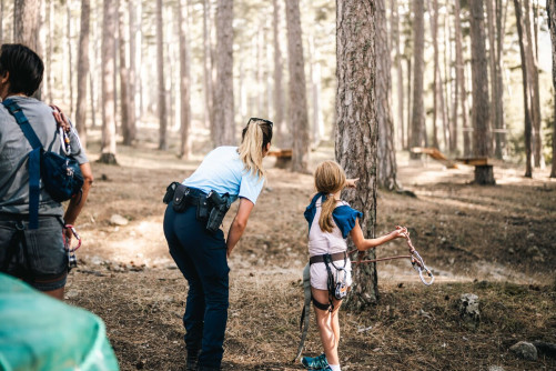 Une femme gendarme, aux cheveux blonds coiffés en queue de cheval, se tient debout, dos à l'objectif, aux côtés d'une petite fille, qui lui désigne un point avec l'index de sa main gauche, dans une forêt