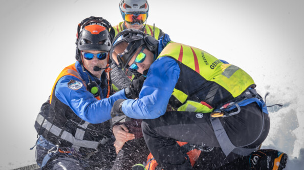 Trois gendarmes secouristes du PGHM protègent une victime de l'envolée de la neige dû à l'arrivée de l'hélicoptère.