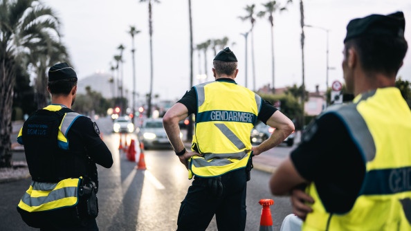 Des gendarmes vêtus d'une chasuble jaune, sont debout, au bord d'une route sur laquelle circulent des véhicules. Des plots oranges sont disposés au sol.