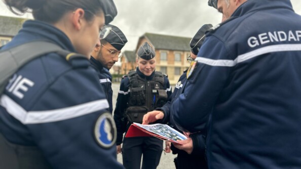 Un groupe de cinq gendarmes - deux femmes et trois hommes - en tenue, se tiennent debout dans la rue, en plein jour. L'un d'eux s'exprime, un cahier &agrave; la main. On aper&ccedil;oit en arri&egrave;re plan des arbres et la fa&ccedil;ade d'un b&acirc;timent en brique rouge.