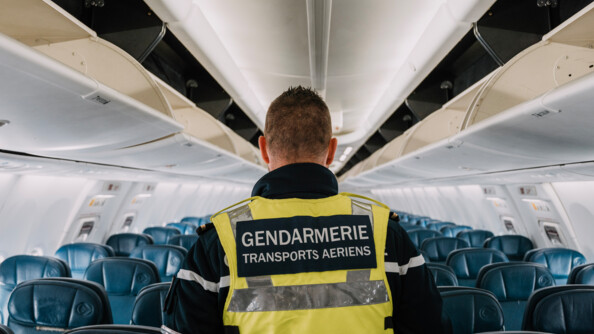 Un gendarme revêtant un gilet jaune portant la mention "Gendarmerie Transports aériens", que l'on voit de dos, se déplace dans l'allée centrale d'un avion de ligne, dont tous les sièges (de couleur bleue) sont vides.