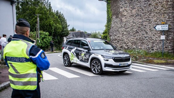 Un gendarme de dos sécurisant le passage du Tour dans un village, tandis qu'un véhicule de l'organisation passe devant lui.