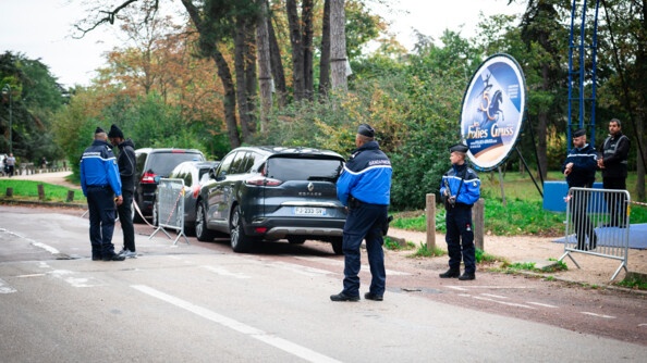 Sur une route, au milieu des bois, qautre gendarmes et un agent de sécuritté se tiennent en faction. L'un deux échange avec un homme vêtu d'un jogging noir et de baskets. Trois véhicules sont stationnés l'un derrière l'autre. On aperçoit en second plan un panneau circulaire sur lequel est inscrit Les Folies Gruss et figurent un cheval et une danseuse