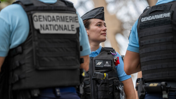 Une jeune gendarme se tient deboit face à l'objectif. Elle regarde su rle côté. Elle est entourée de deux collègues gendarmes, qui tournent le dos à l'objectif. Tous trois sont équipés d'un gilet pare-balles. Derrière eux, on devine la structure métallique de la Tour Eiffel.