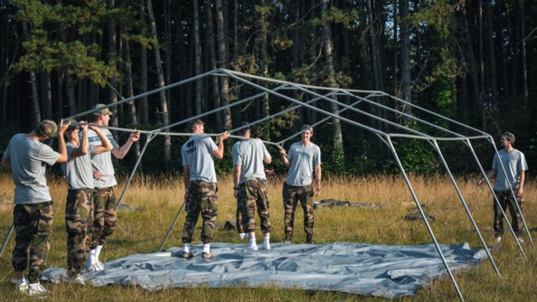 Sept jeunes hommes, en treillis, baskets et tee-shirts gris, montent une structure de tente dans un champ, en lisière de bois.