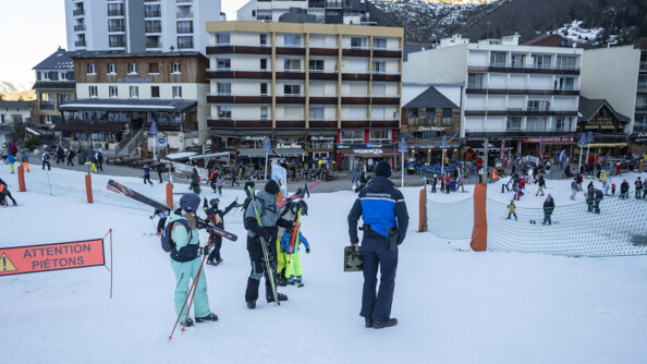 Un gendarme de dos près de skieurs à sa gauche, sur une pente enneigée juse au-dessus d'une station avec des commerces, restaurants, immeubles et touristes