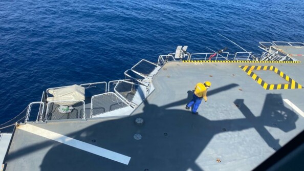 Vue sur la mer et sur le pont du porte hélicoptères Mistral, où se projette l'ombre de l'hélicoptère de la gendarmerie nationale. Sur le pont, un agent de piste vêtu d'une chasuble et d'un casque jaunes, vu de dos.