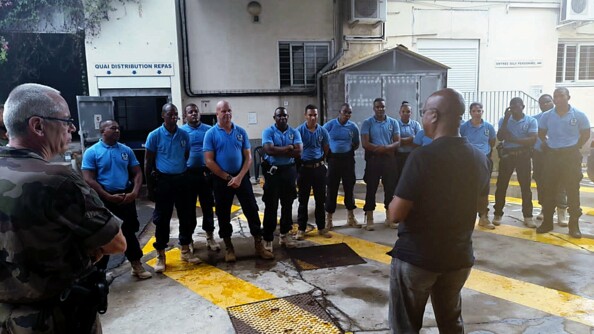 Deux hommes de dos, l'un en treillis kaki et l'autre en civil, s'adressent à une rangée de policiers seychellois vêtus de pantalons de treillis noirs et de polos bleus.