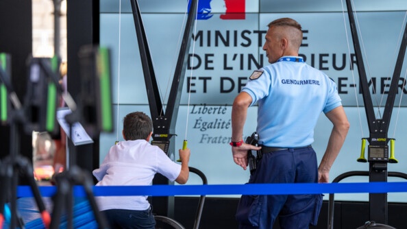 Un enfant que l'on voit de dos joue sur une machine suspendu par les bras. A ses côtés se trouve un gendarme, de dos également. On devine l'inscription ministère de l'intérieur et des outre mer sur le mur face à eux.