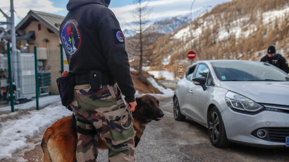 Un maître de chien et son malinois devat un véhicule gros contrôlé par un deuxième gendarme en arrière-plan, ainsi que la montagne semi-enneigée
