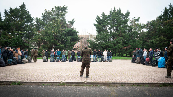Sur une place d'arme, les futurs personnels du Groupe d’Observation et de Surveillance se préparent à débuter leur formation. Entourés d'arbres, ils sont positionnés en carré devant leur formateur.