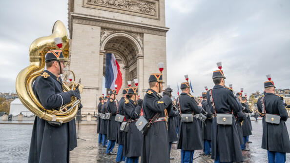 Des musiciens de la Garde républicaine en tenue, vêtus d'un long manteau noir, pantalon bleu marine, et du traditionnel chapeau à plumes, jouent divers instruments de musique sur le sol pavé de la place Charles de Gaulle à Paris. Derrière eux, se trouve l'Aec de Triomphe. Le ciel est gris.