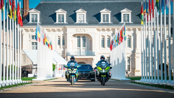 Dans une large allée, devant un château, une berline noire avance, précédée de deux gendarmes motocyclistes. De nombreux drapeaux de différents pays, non hissés, jalonnent les bords de l'allée.