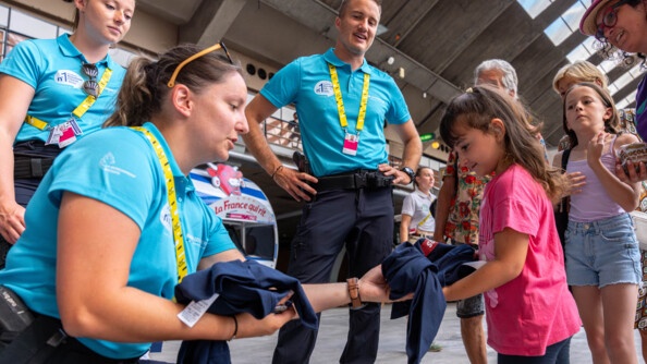 Une jeune femme gendarme, accroupie, tend un t-shirt bleu marine, qu'elle offre à une fillette vêtue d'un t-shirt rose. Derrière eux se tiennent d'autres personnes disposées en cercle derrière elles, dont deux gendarmes, et une seconde fillette.