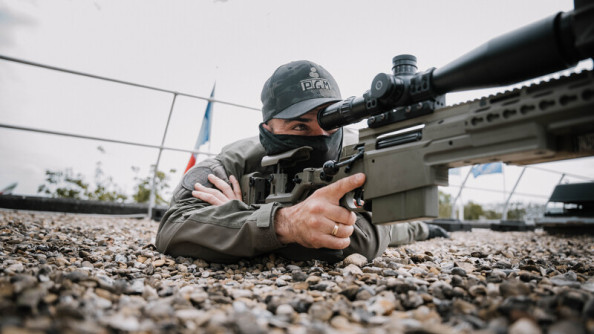 Un gendarme tireur d'élite est en position, couché à plat ventre sur un toit, le doigt sur la gachette de son fusll. On aperçoit le drapeau français en arrière plan.