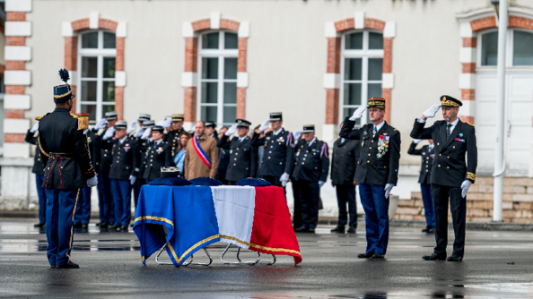 Des militaires saluent un cercueil recouvert du drapeau français. Au milieu d'eux, se trouve un élu, vêtu d'un manteau beige et de l'écharpe tricolore