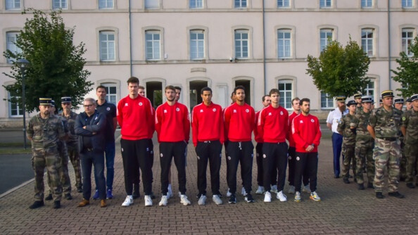 Au centre de la photo, douze jeunes hommes en bas de jogging noir, sweat rouge, répartis sur deux rangées. A gauche, deux hommes plus âgés en tenue bleue et trois militaires en kaki, l'un derrière l'autre. A droite, des militaires en kaki.