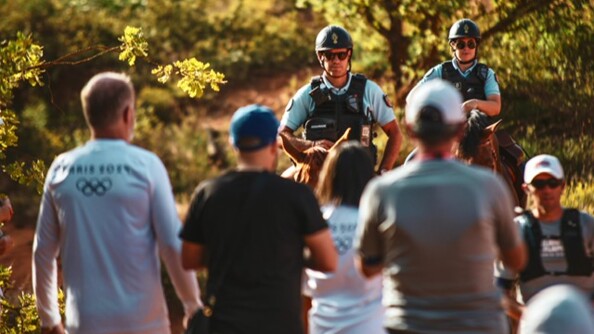 Au premier plan, quatre personnes de dos, dont deux portant un tee-shirt blanc siglé Paris 2024. Au second plan, à droite, un homme vu de face portant une casquette blanche et des lunettes de soleil, suivi par deux cavaliers de la gendarmerie sur leur monture, dont on n'aperçoit que les oreilles et le sommet de la tête.