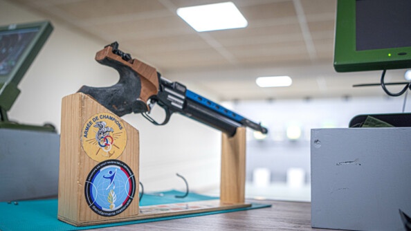 Photo de l'arme de tir posée sur un socle en bois. sur celui-ci on peut voir deux rondaches dont un de l'armée des champions, et l'autre des sportifs de haut niveau de la défense.