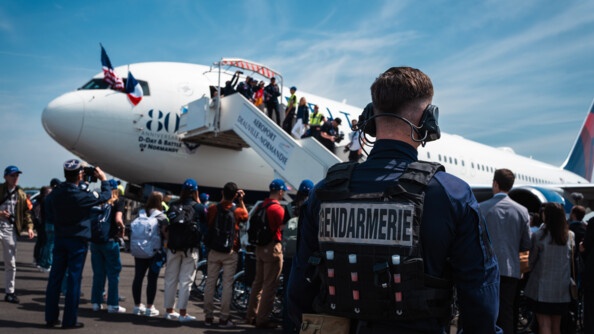 Un gendarme de dos à droite sur le tarmac où une foule nombreuses se presse à la descente d'un avion blanc siglé aux 80 ans du débarquement
