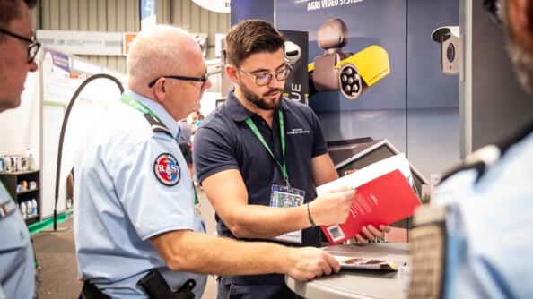 Un gendarme référent sûreté de profil droit échnage avec un homme en polo bleu marine qui lui montre une brochure. Devant eux en fond un kakemono pour de la video surveillance