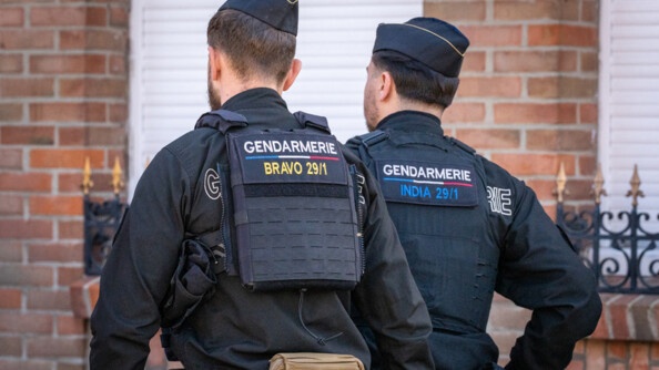 La photo repr&eacute;sente deux gendarmes avan&ccedil;ant dans la rue au c&ocirc;t&eacute;s des carnavaleux, de dos, devant une maison en briques rouges.
